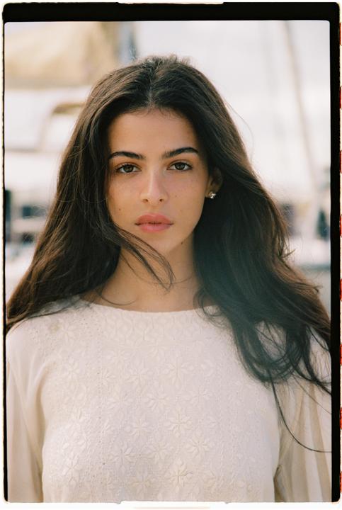 A woman with long dark hair and a white embroidered top looks directly at the viewer.