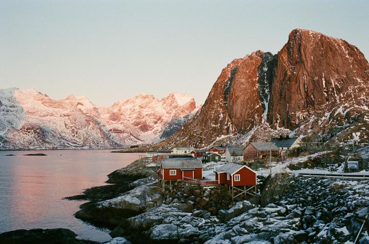 A small village with red and white houses sits on a rocky, snow-covered shore next to a body of water, with large, snow-capped mountains in the background.