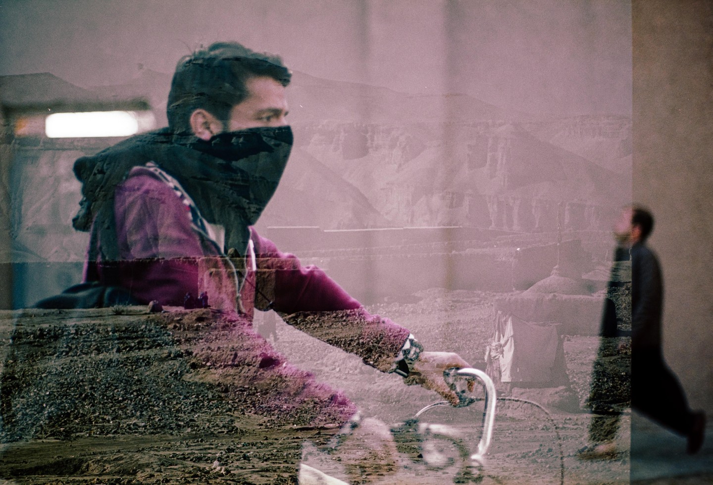 A man with a covered face is riding a bicycle, with a faint image of a mountain landscape and another man walking in the background.