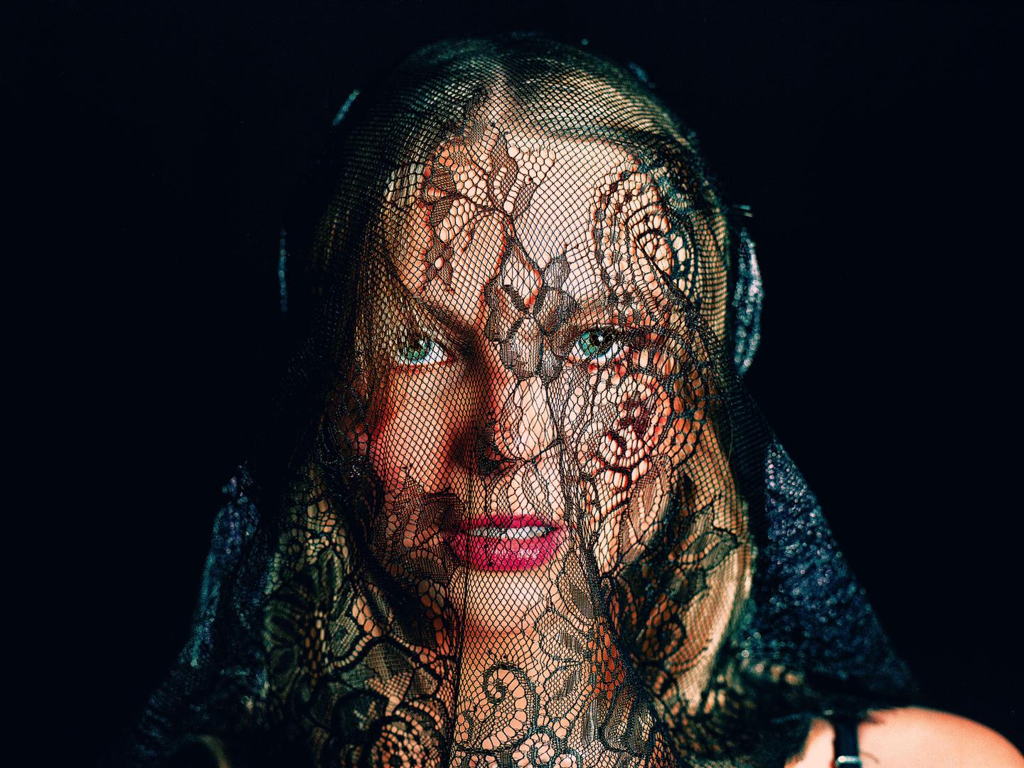 A woman with red lipstick and green eyes looks directly at the viewer through a black lace veil.