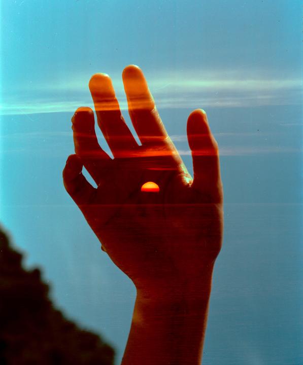 A double exposure shows a hand superimposed over a sunset with a blue sky and ocean.