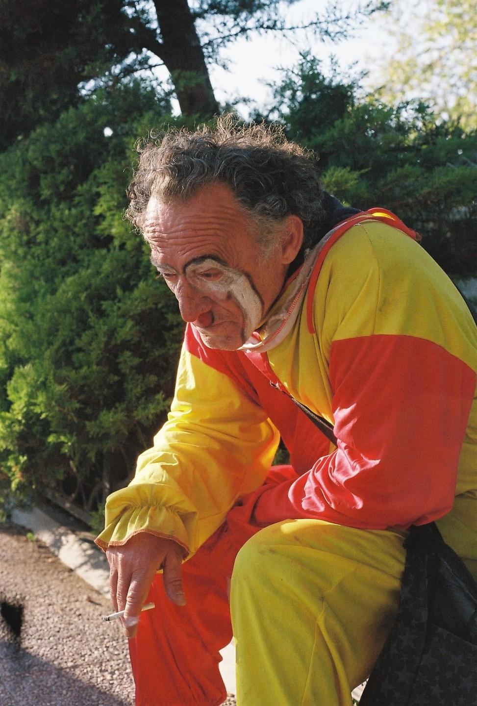A clown in a yellow and red costume sits outdoors, smoking a cigarette.