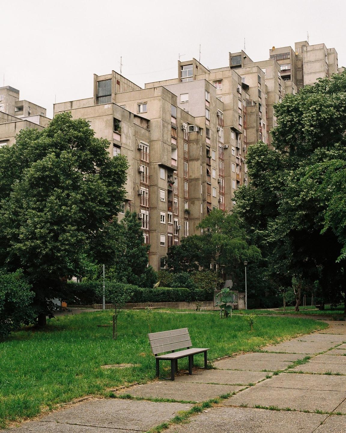 A concrete apartment building rises behind a park bench and a path.