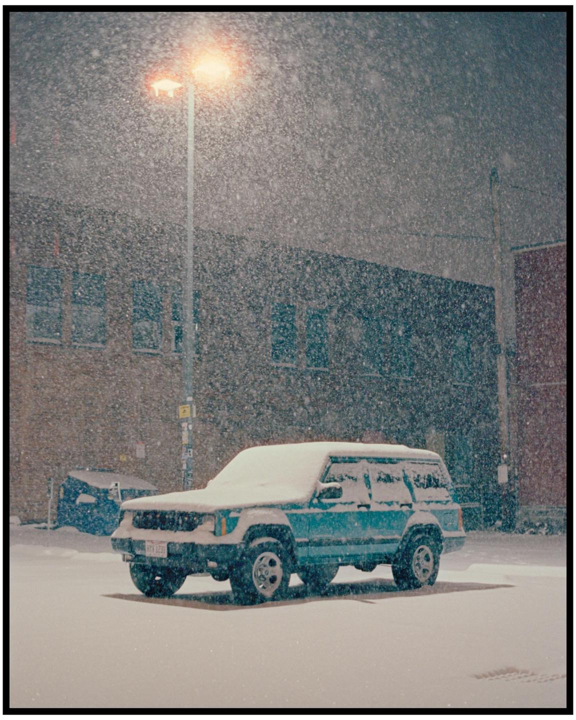 A blue sport utility vehicle is parked on a snow-covered street under a street light during a snowstorm at night.