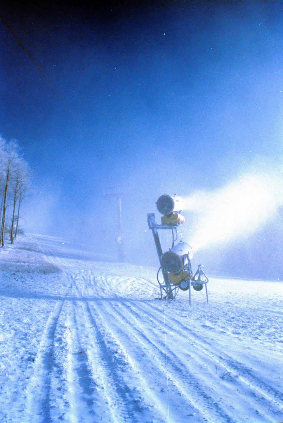A snow cannon sprays snow onto a ski slope at night under a dark blue sky.