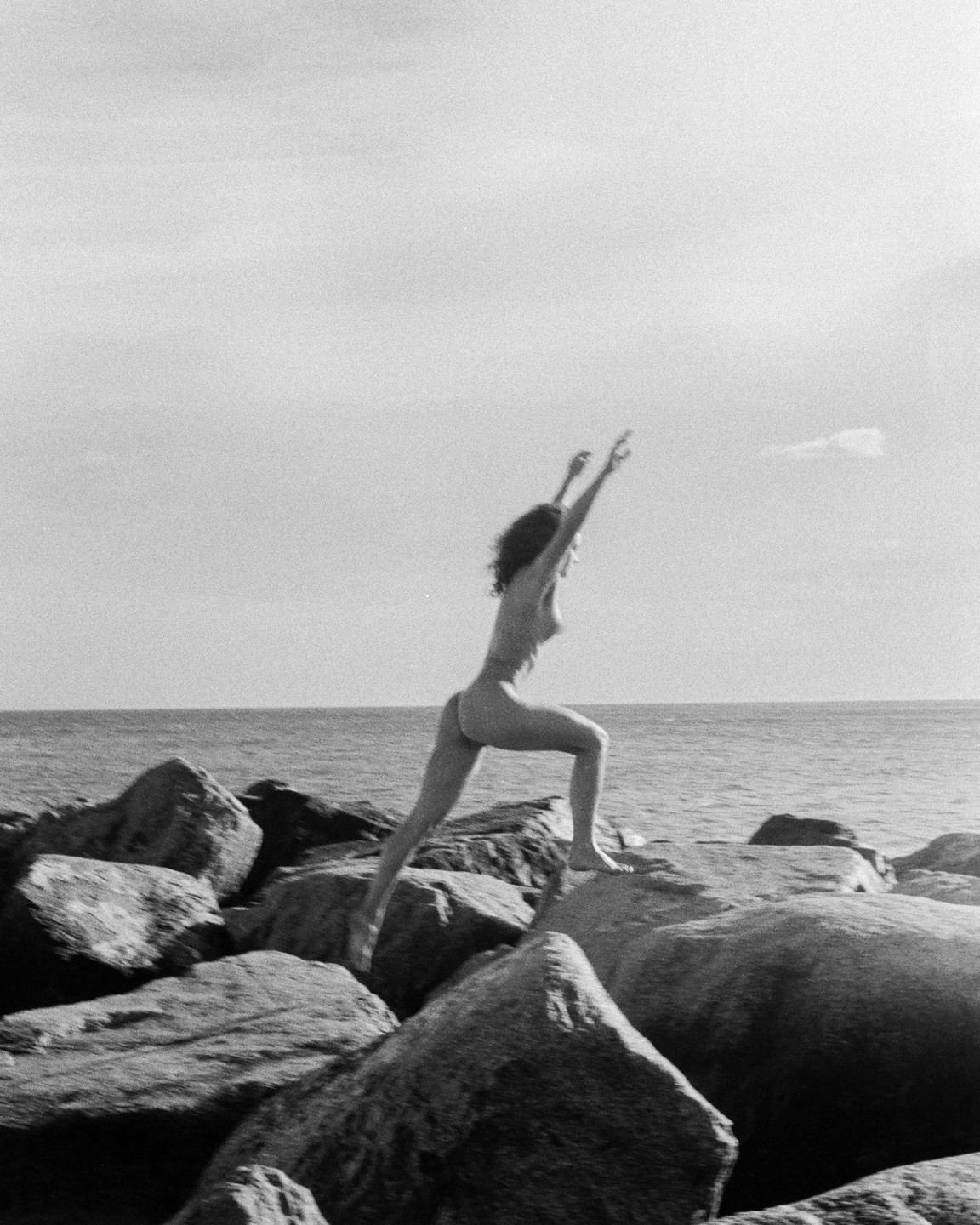 A woman in a swimsuit jumps between rocks by the ocean.