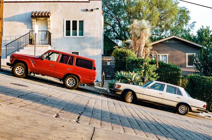 Two cars are parked on a steep, paved street in front of houses and lush vegetation.