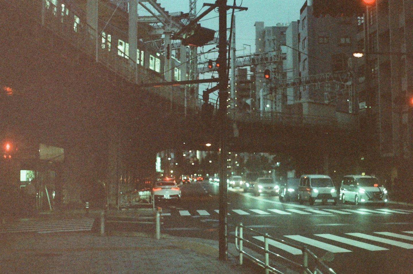 A street at night shows cars driving on a wet road under an overpass with buildings and traffic lights visible.