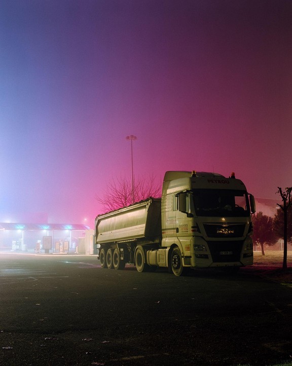 A large truck is parked in a lot with a gas station in the background, all shrouded in colorful fog.