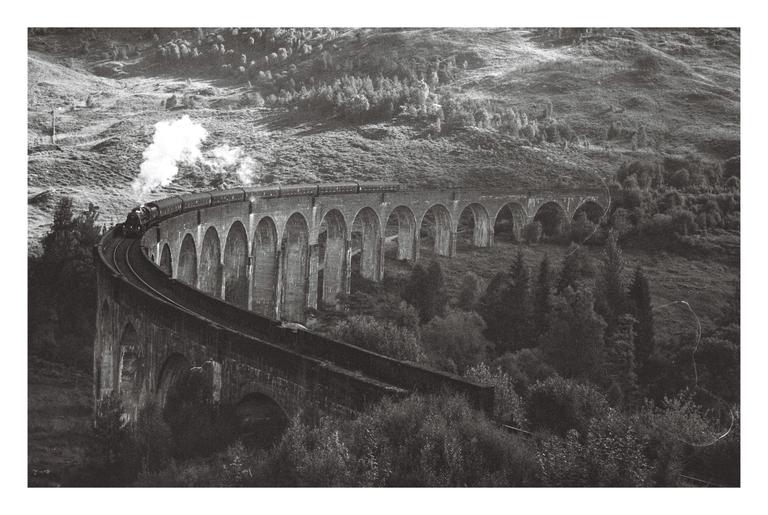 A train with a plume of smoke travels across a long, arched viaduct in a hilly, forested landscape.