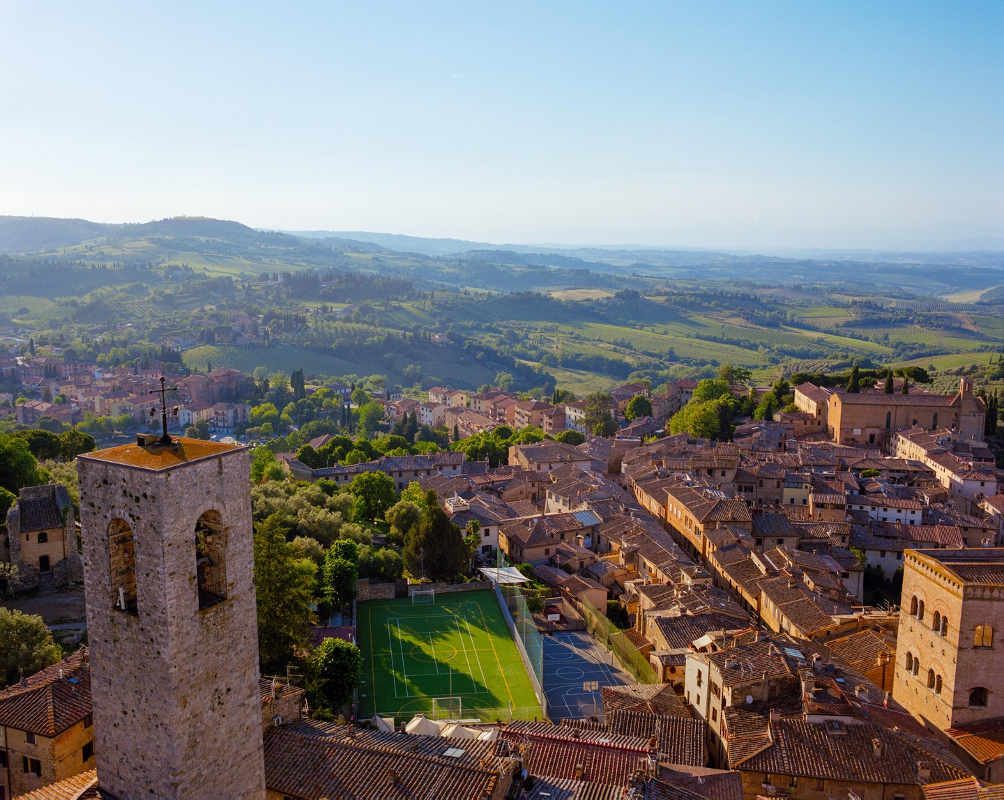 A high-angle view shows a town with a bell tower, buildings with terracotta roofs, a sports field, and rolling hills in the background.