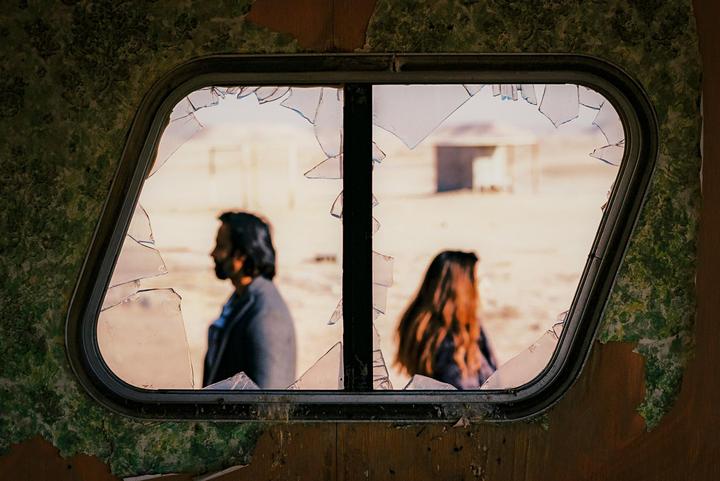 A man and a woman are visible through a broken window with peeling wallpaper.