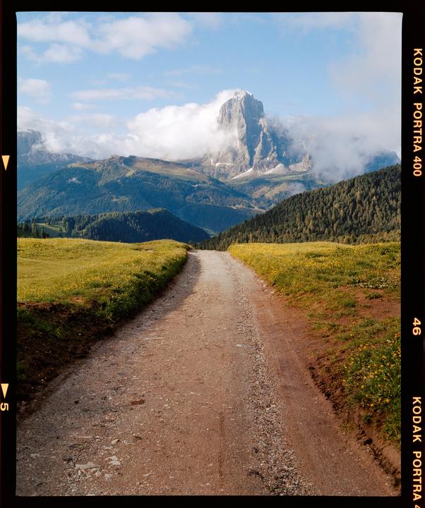 A dirt road leads through a green field towards a large mountain partially obscured by clouds.