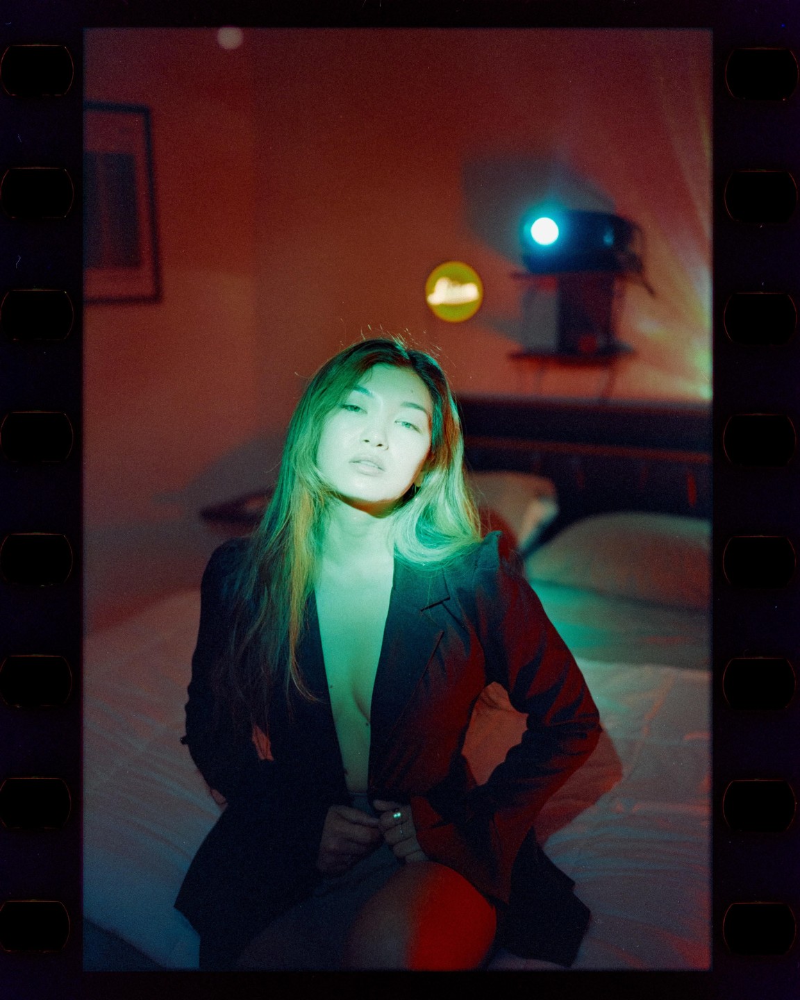 A woman sits on a bed in a dark room with a projector light shining on her.