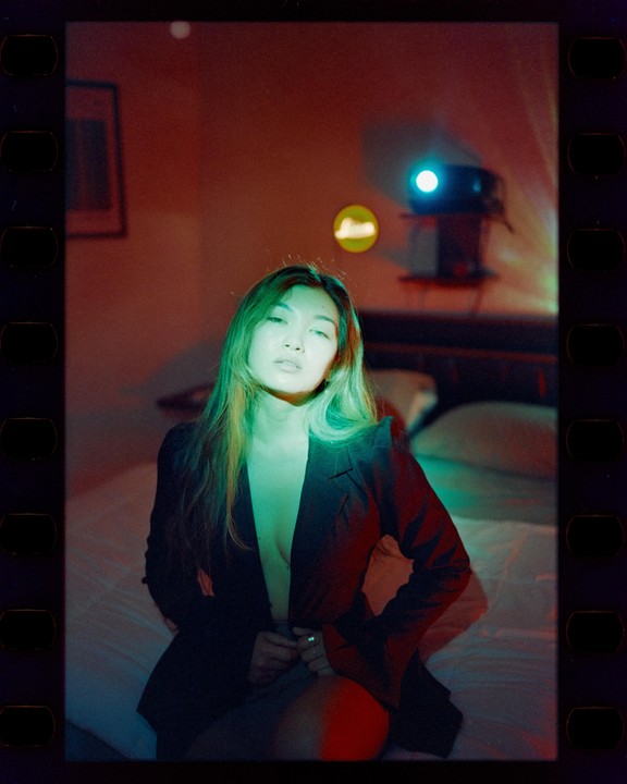A woman sits on a bed in a dark room with a projector light shining on her.