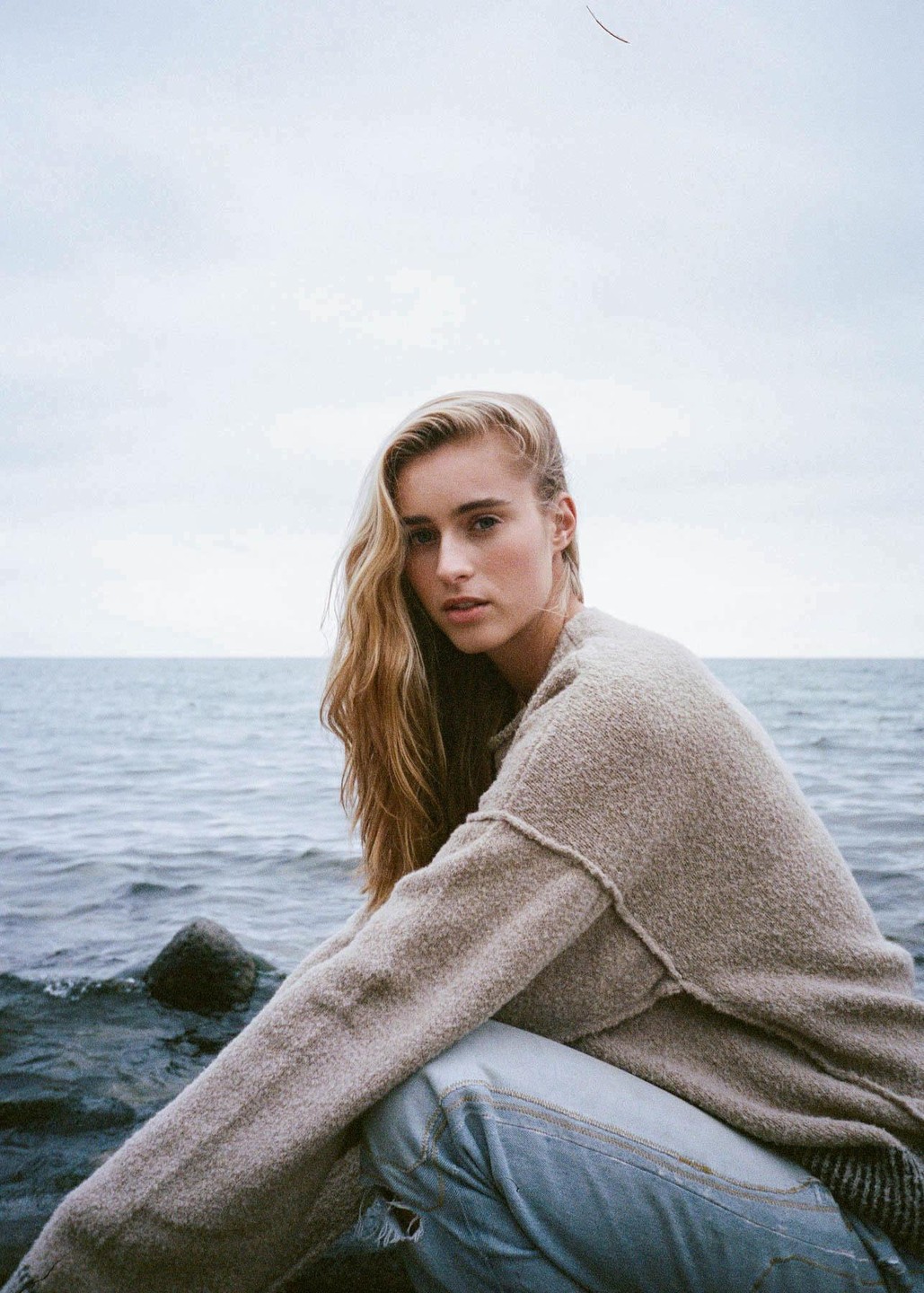 A woman with long blonde hair crouches by the water, looking directly at the viewer.