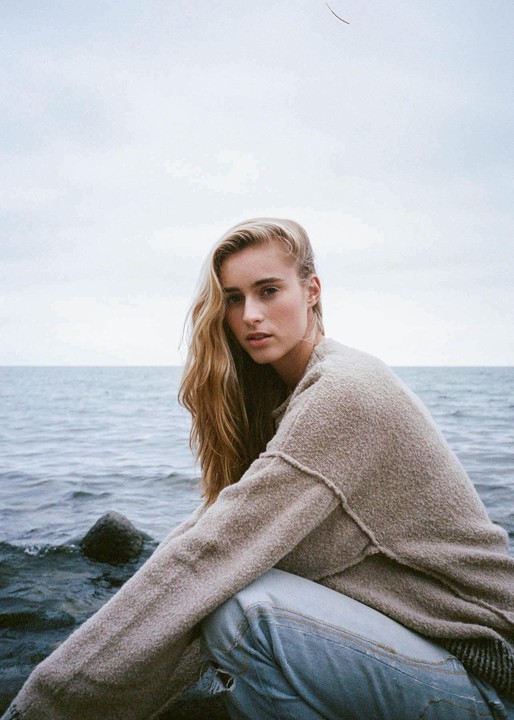 A woman with long blonde hair crouches by the water, looking directly at the viewer.