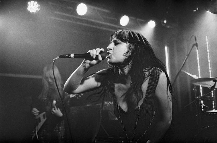 A woman sings into a microphone on a stage with a drummer and guitarist in the background.