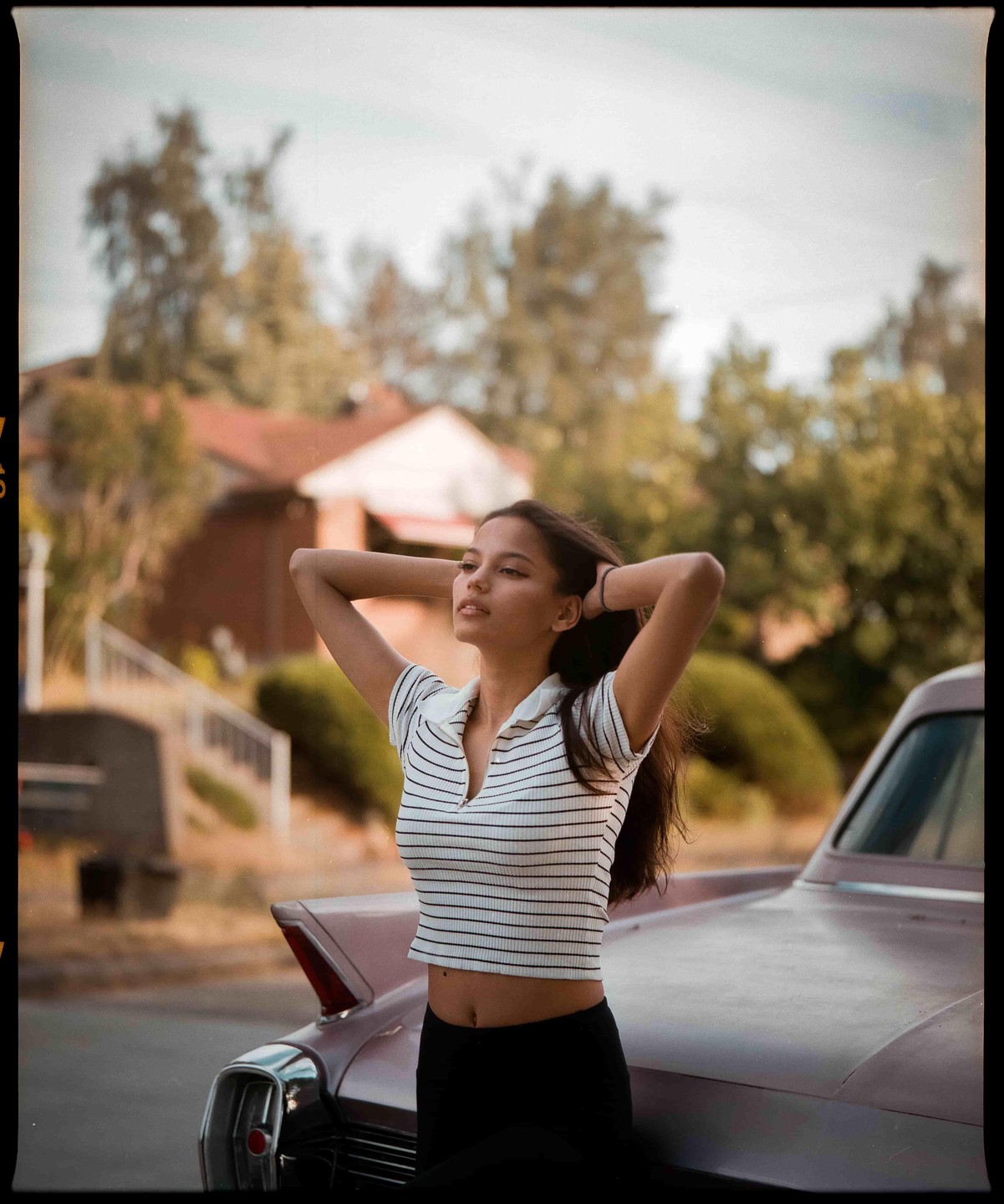 A woman with her hands behind her head leans against a classic car on a street.
