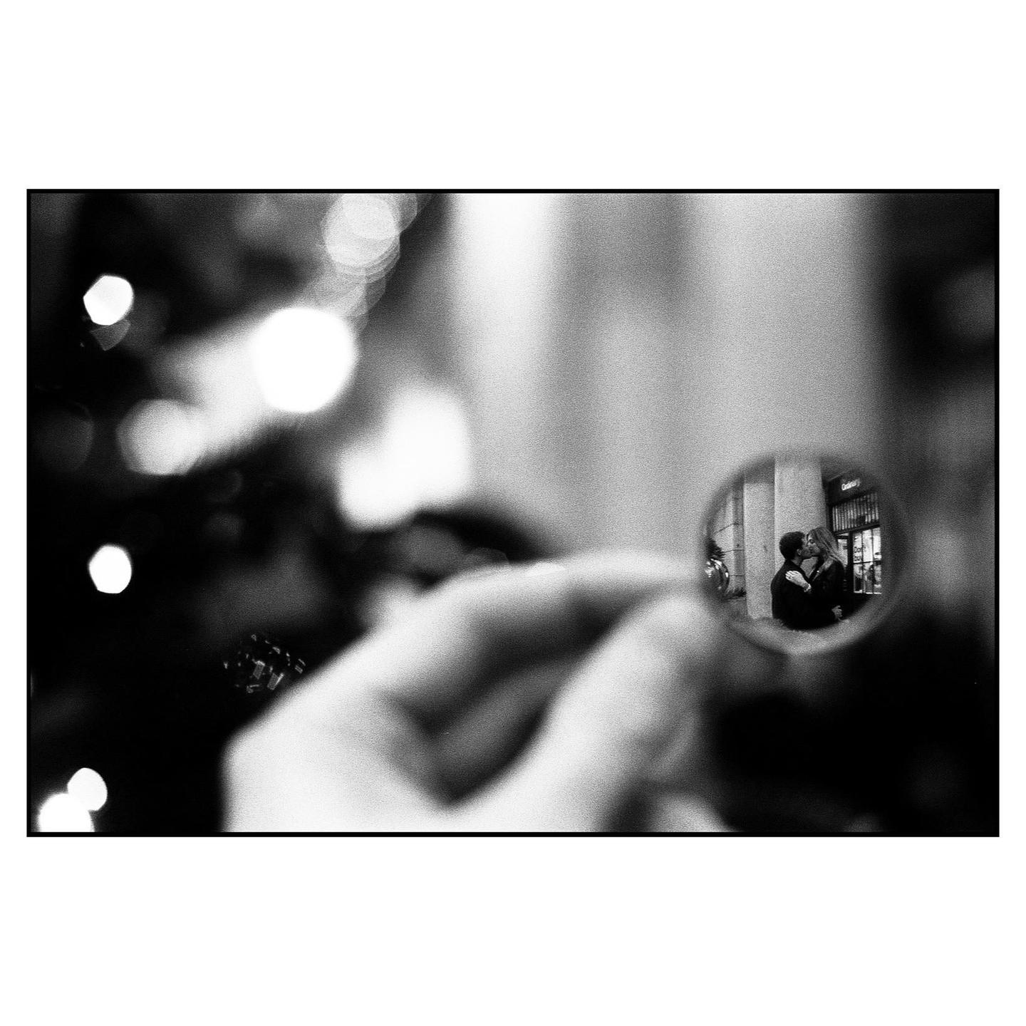 A hand holds a small circular mirror reflecting a couple kissing on a street.