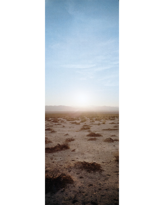 The sun sets over a vast desert landscape with sparse vegetation and distant mountains.