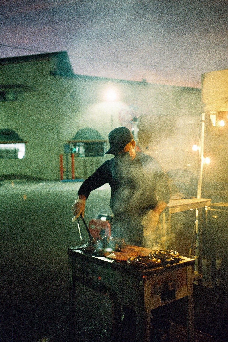 A man wearing a baseball cap and gloves cooks meat on a grill at night, with smoke rising from the food.