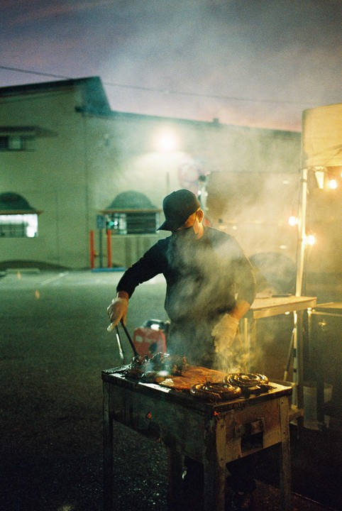 A man wearing a baseball cap and gloves cooks meat on a grill at night, with smoke rising from the food.