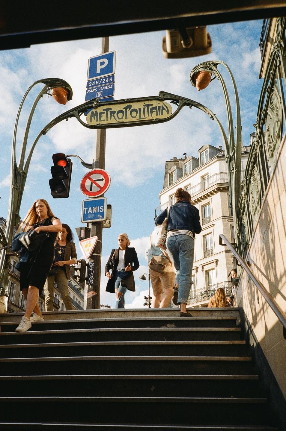 People walk up and down the steps of a Paris Metro station entrance on a sunny day.