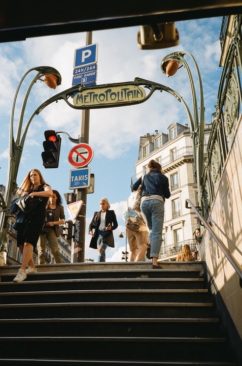 People walk up and down the steps of a Paris Metro station entrance on a sunny day.