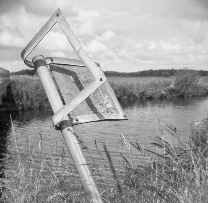 A warped metal sign stands in tall grass beside a body of water with a distant treeline under a cloudy sky.
