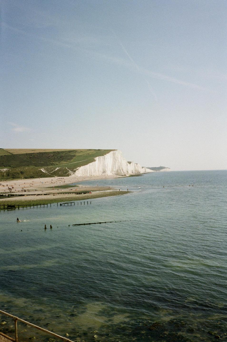 A wide view shows the Seven Sisters cliffs along the coast with people on the beach and in the water.
