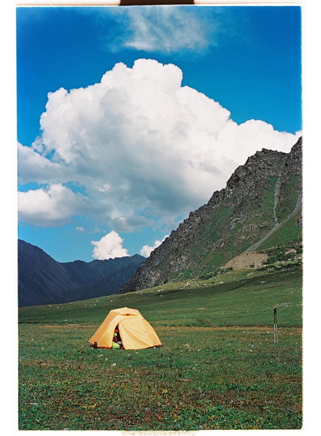 A yellow tent sits in a green field with mountains and a blue sky with clouds in the background.