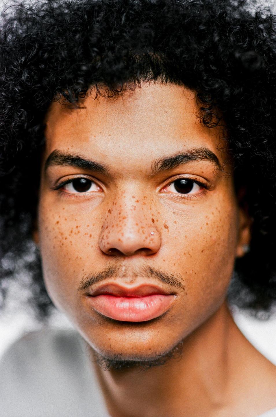 A man with curly hair, freckles, and a nose piercing looks directly at the viewer.