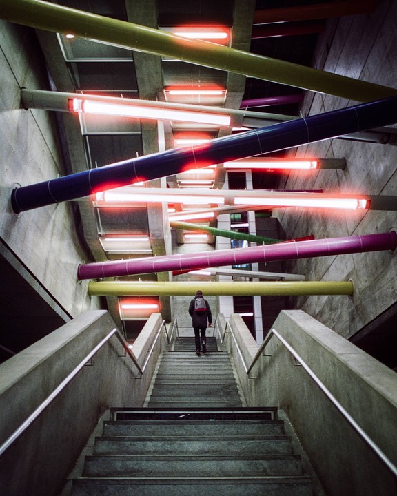 A person ascends a concrete staircase in a subway station with colorful pipes and red neon lights.