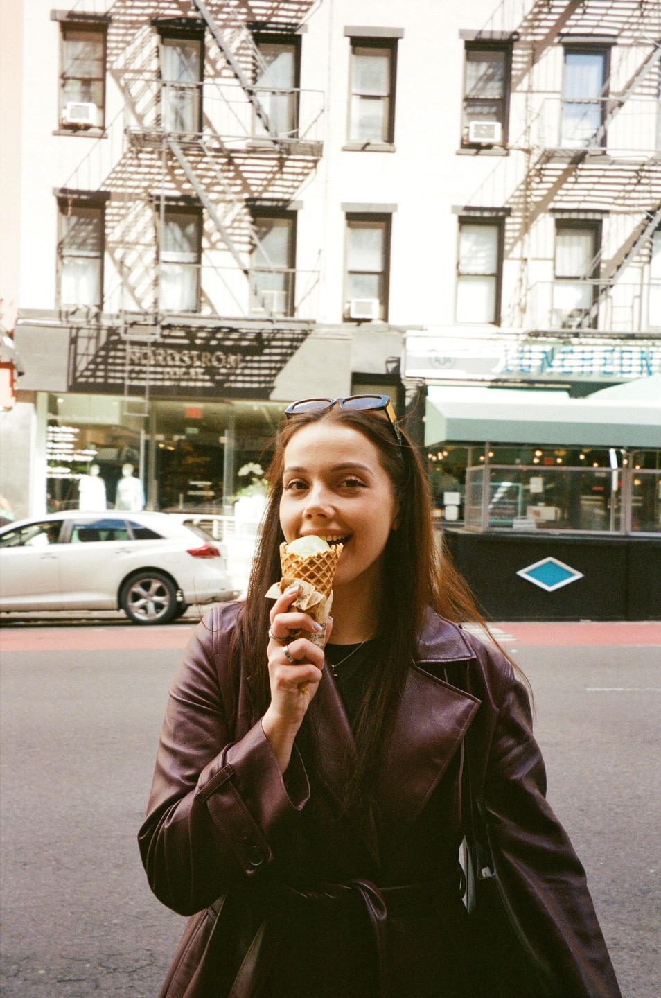 A woman in a brown coat eats an ice cream cone on a city street.
