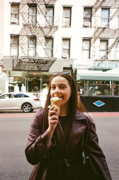 A woman in a brown coat eats an ice cream cone on a city street.