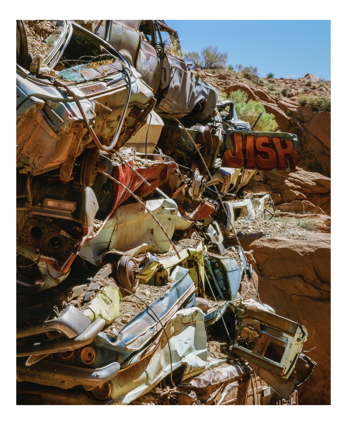A pile of rusted, crushed cars forms a retaining wall in a desert landscape, with the word "JISH" spray-painted on one of the car doors.