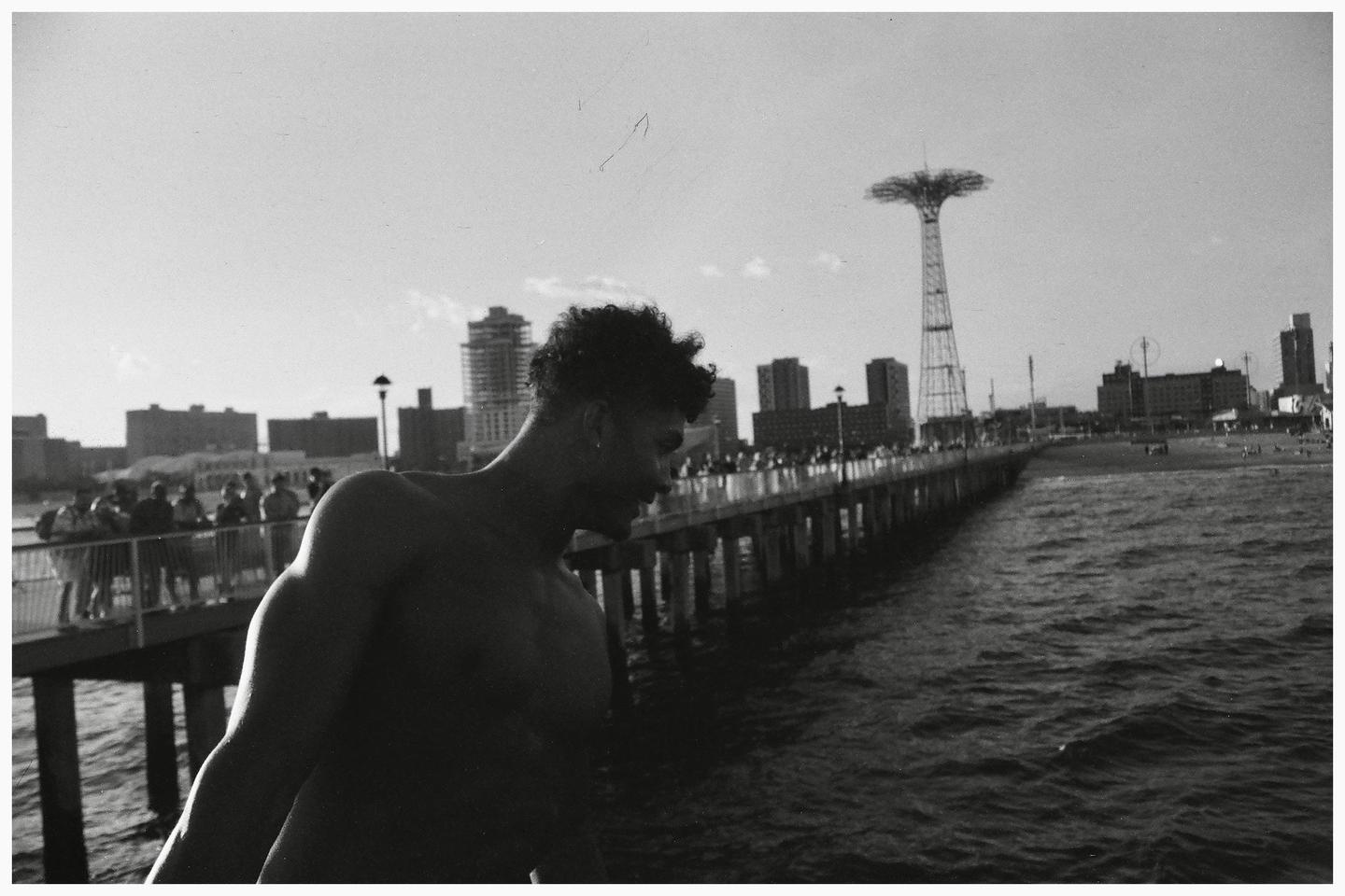 A shirtless man stands on a pier with a city skyline and a parachute jump tower in the background.
