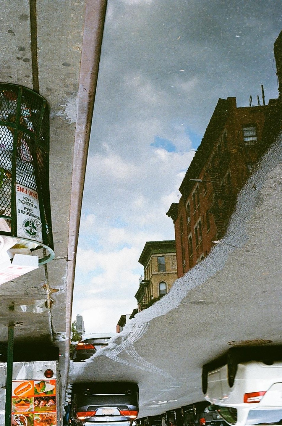 A street scene is reflected in a puddle, showing buildings, cars, and a food cart.