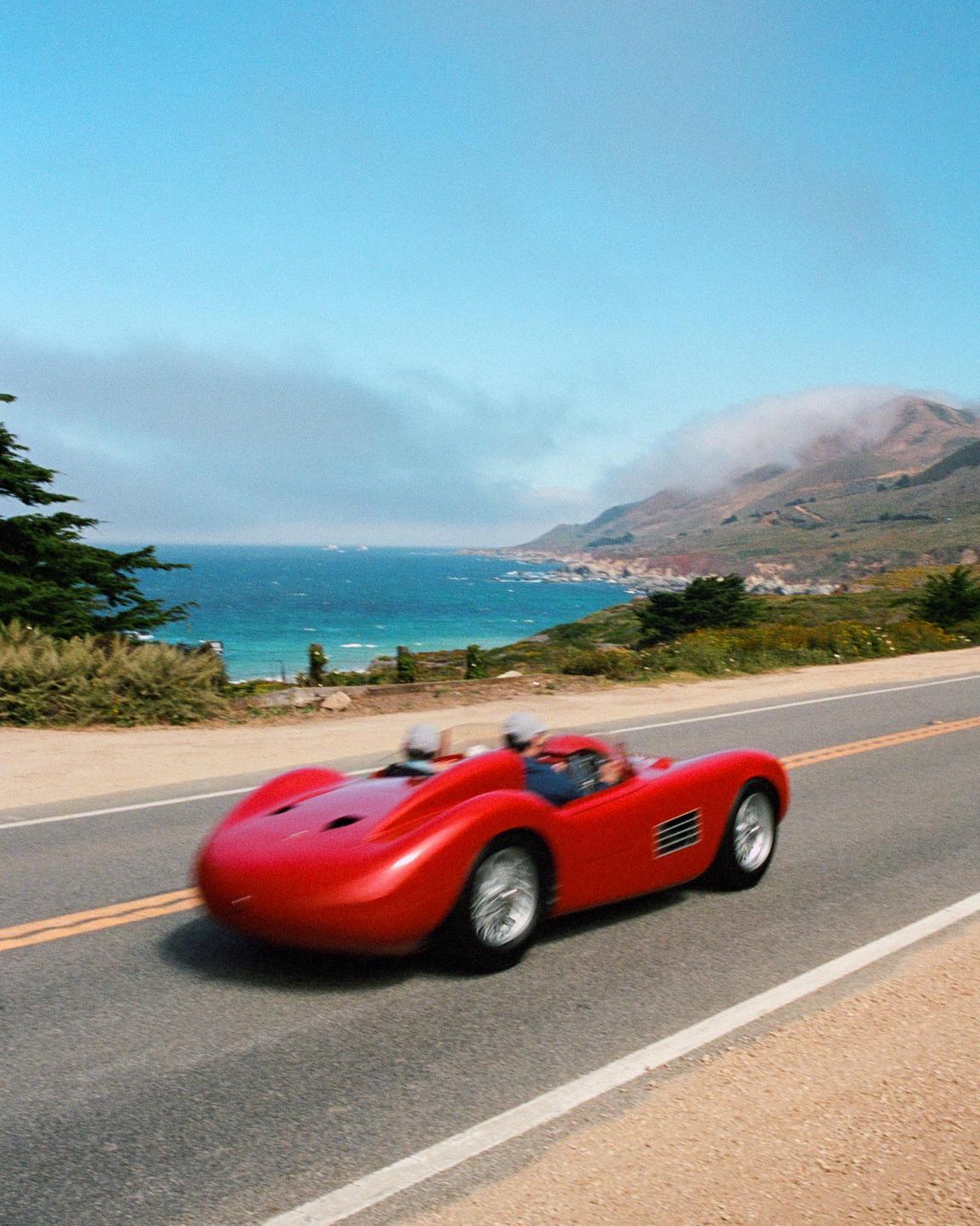 A red vintage race car drives along a coastal road with a blurred background of the ocean and mountains.