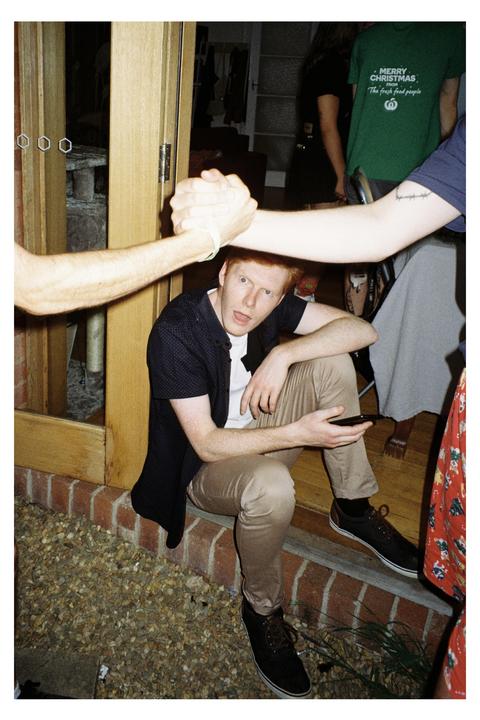 A man with red hair sits on a brick ledge while two arms shake hands above his head.