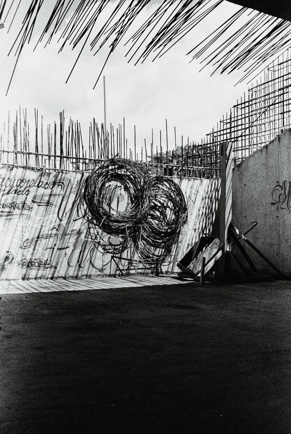 Two large coils of barbed wire rest against a graffiti-covered wall with rebar and scaffolding visible behind it.