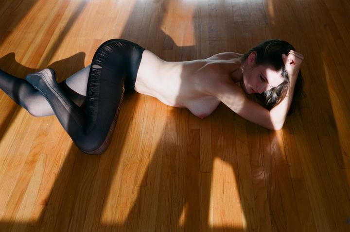 A woman in torn leggings lies on a wooden floor with sunlight and shadow.