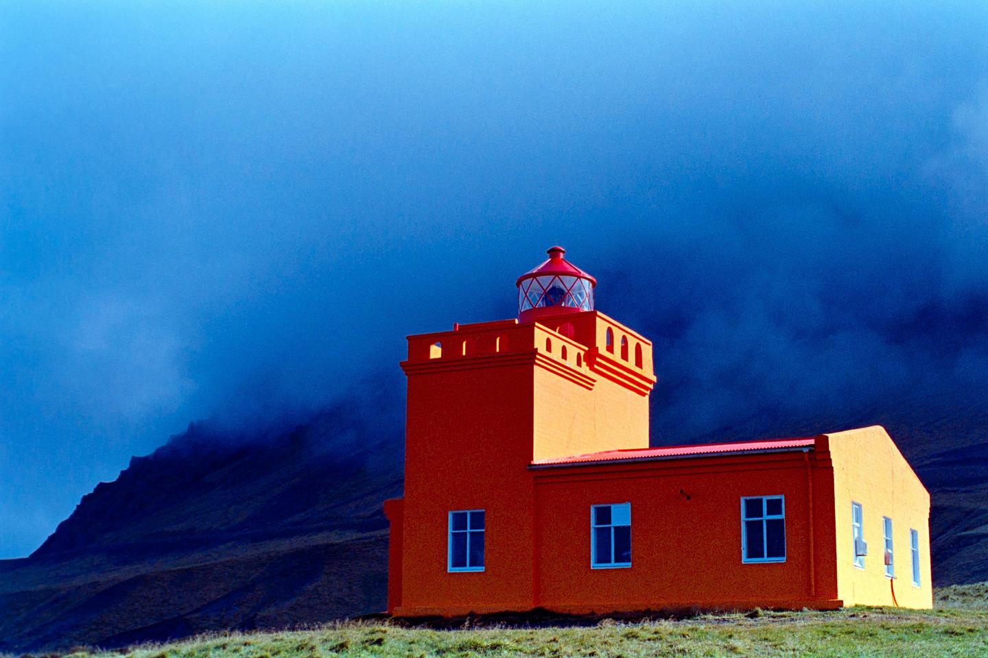 An orange lighthouse stands on a grassy hill with a dark blue, cloudy sky and mountain in the background.