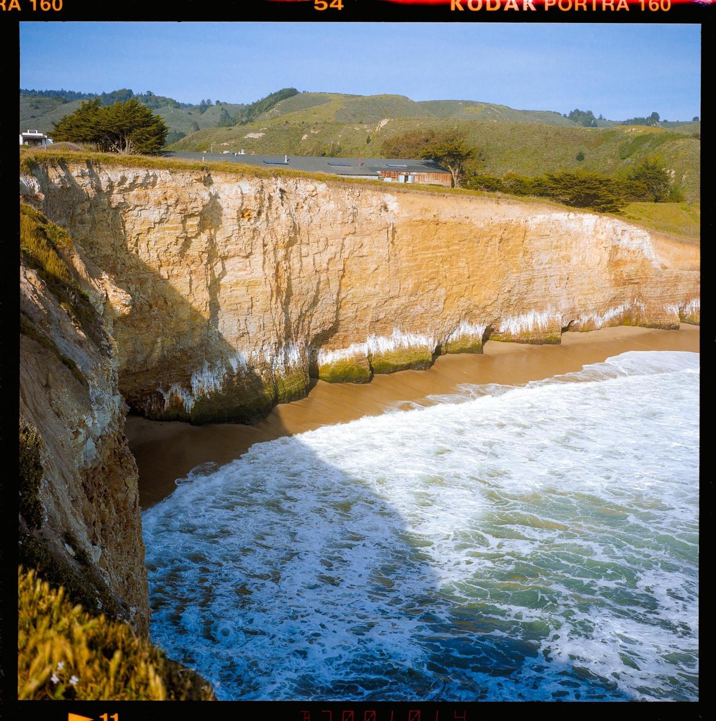 Waves crash on a sandy beach at the base of a tall cliff with green hills and a building in the distance.