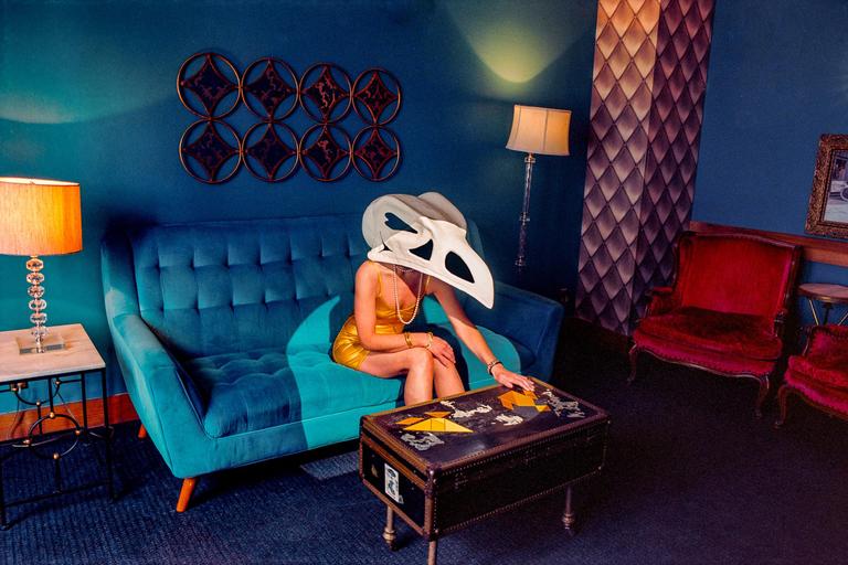 A woman wearing a bird skull mask sits on a blue sofa in a room with blue walls and red chairs.