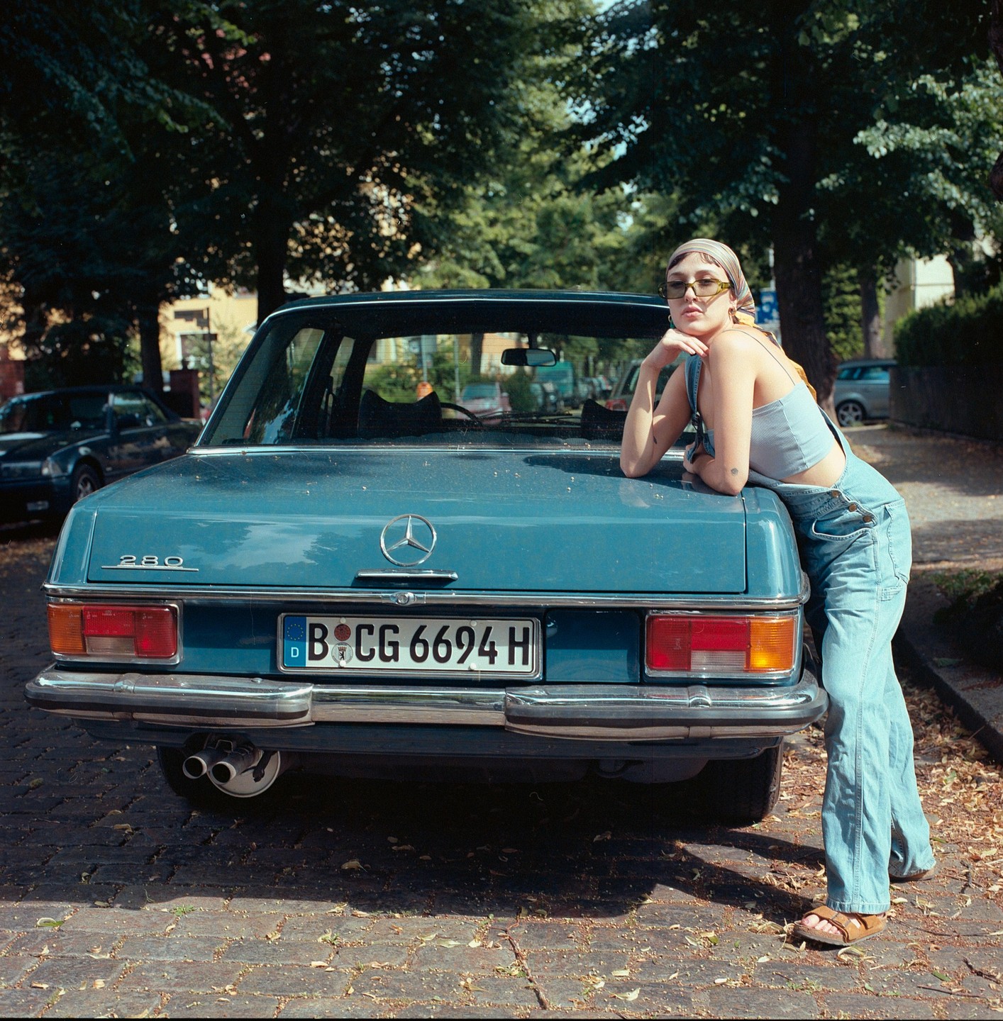 A woman in overalls and sunglasses leans on the trunk of a blue car parked on a street.