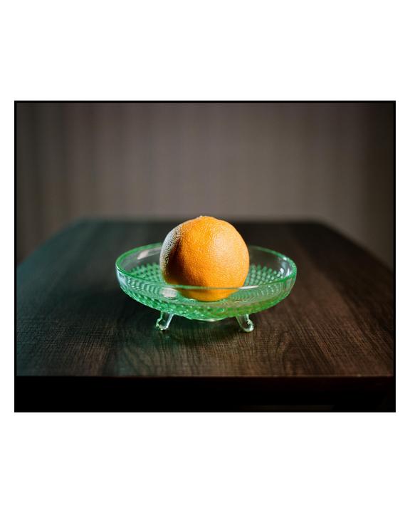 An orange rests in a green glass bowl on a wooden table.