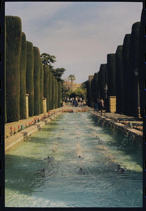 A long, narrow water feature with multiple fountains is flanked by tall, manicured hedges and a path with people in the distance.