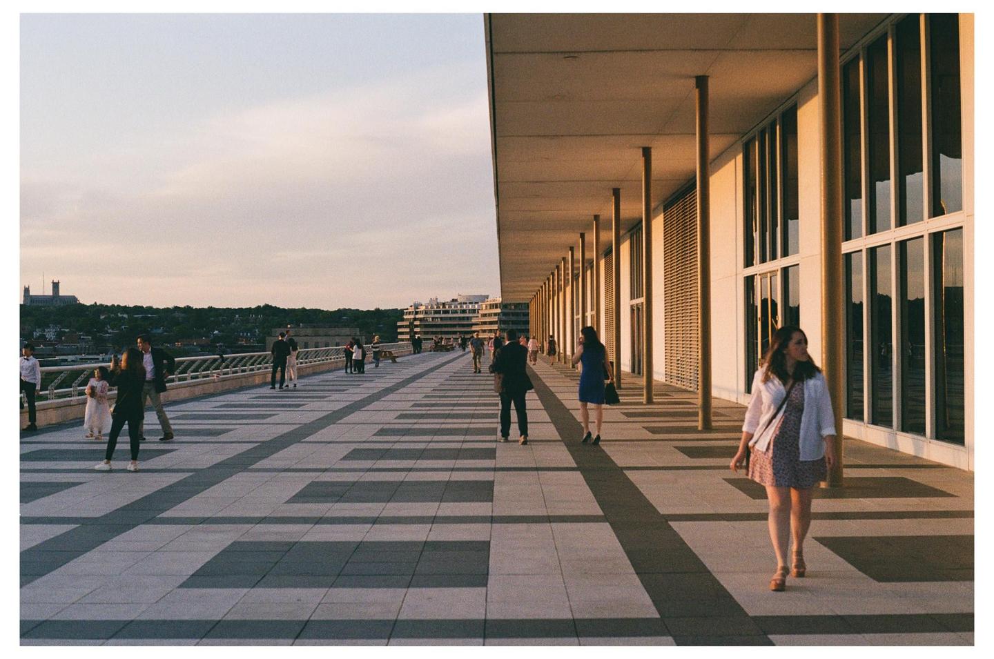 A woman walks along a long outdoor walkway next to a building with many windows, while other people gather in the distance.